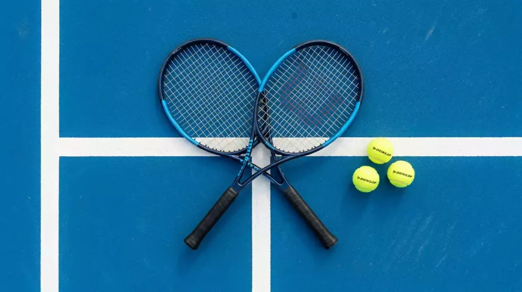 Raquetas de tenis cruzadas sobre una pista dura con pelotas de tenis y la red de fondo